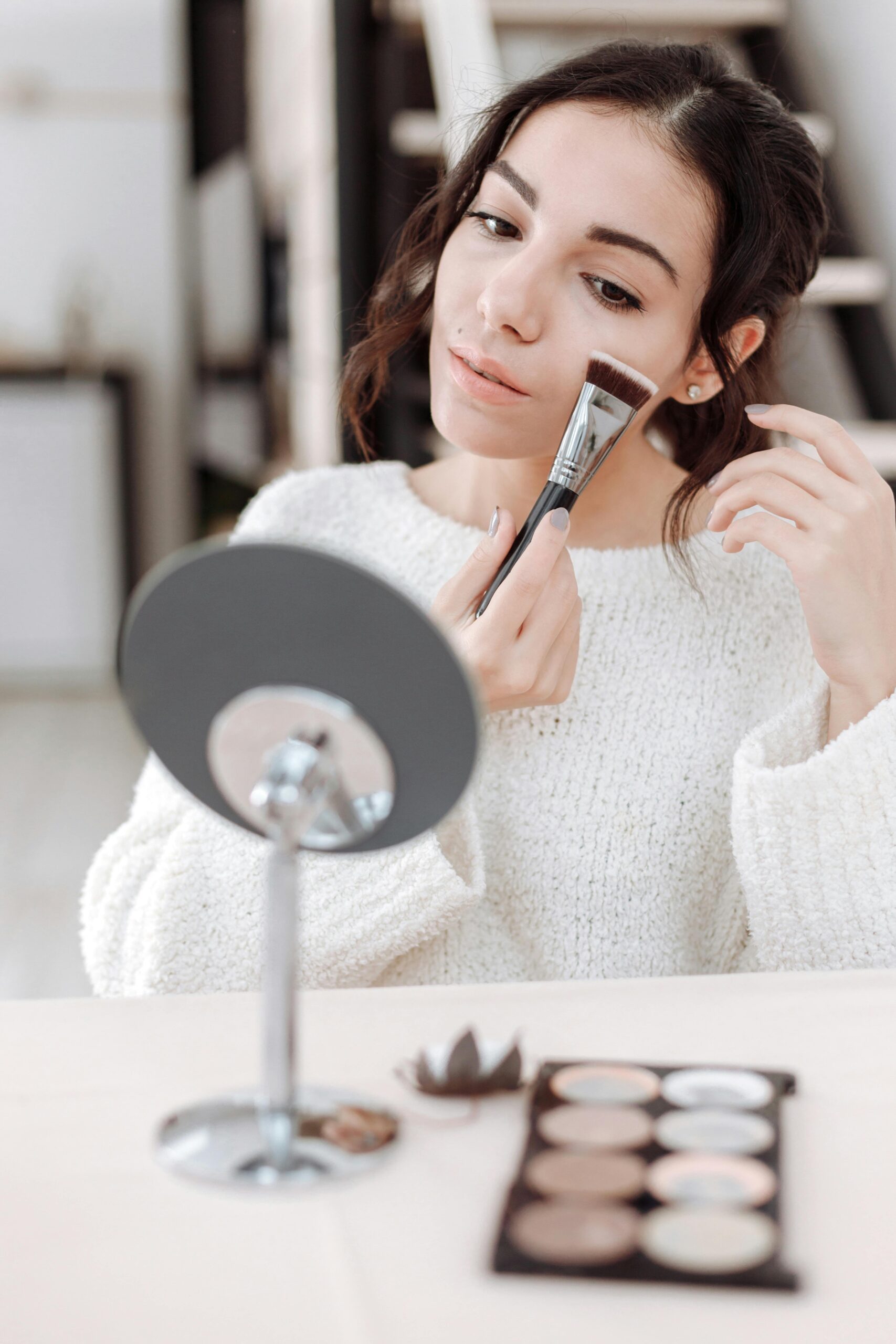 Woman applying face makeup with a brush while looking in a mirror.